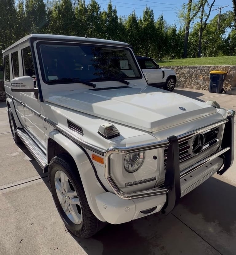 White Mercedes-Benz G-Class SUV with front grille guard parked in lot with trees in background