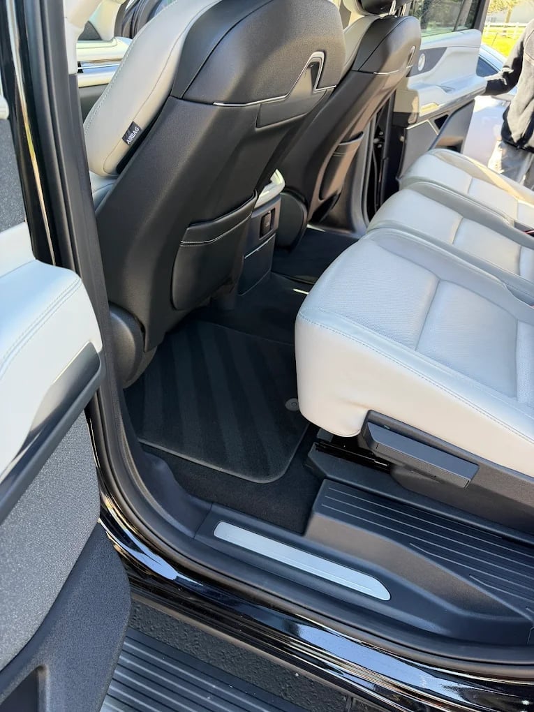 Car interior view showing front and rear seats with white upholstery, black floor mats, and open passenger door
